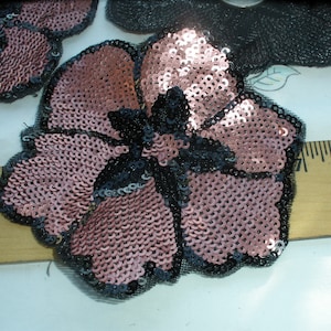 May include: A flower-shaped applique patch covered in shimmering pink sequins with a black sequined center and black mesh trim. The patch is positioned against a wooden ruler, showing it measures approximately 3 inches in width.