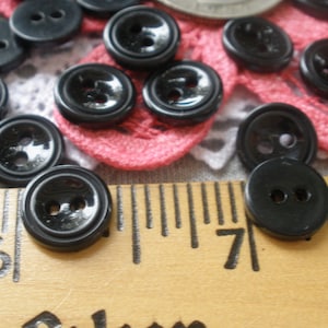 May include: A collection of small, round, black buttons with two holes each. The buttons are scattered on a textured surface with a wooden ruler showing inch measurements. Some buttons are on a pink and white lace doily.