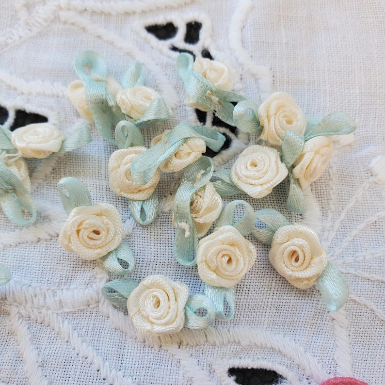 May include: A collection of small, cream-colored satin ribbon roses with light blue ribbon leaves and loops. The roses are scattered on a white, patterned fabric, creating a delicate and feminine aesthetic. Ideal for crafting and embellishments.
