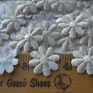 May include: A collection of embroidered, light gray flower appliques. The appliques are in the shape of a daisy with detailed stitching. A wooden ruler with the text "Mother Goose Shoes" is in the foreground.