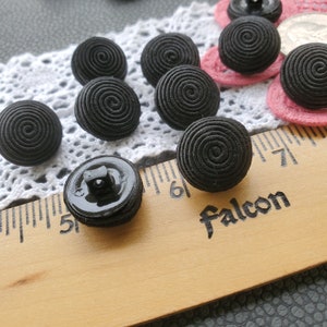 May include: A close-up of 10 black spiral buttons, some with a shiny black finish. The buttons are arranged on a white lace doily and a wooden ruler marked with inches and the word "Falcon".