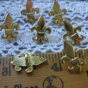 May include: A set of seven gold-colored metal fleur-de-lis buttons. The buttons are arranged on a white lace fabric background. A wooden ruler with inch markings is in the foreground.