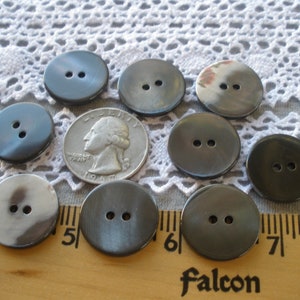 May include: Nine black and gray two-hole buttons with a shiny finish. The buttons are arranged in a semi-circle on a white lace tablecloth. A ruler is visible in the bottom of the image.