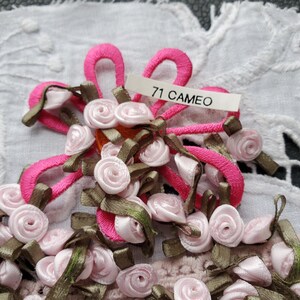 May include: A collection of small, pink ribbon roses with green ribbon accents and bright pink looped ribbon. A tag reads "71 CAMEO". The roses are arranged on a white, textured surface.