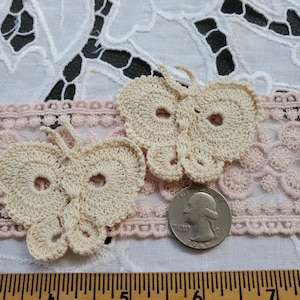 May include: Two crocheted cream-colored butterfly appliques with a hole in the center of each wing. The butterflies are on a white lace background with a pink lace trim.