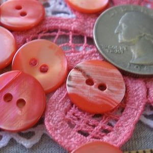 May include: A collection of pink and white shell buttons with two holes each. The buttons are arranged on a pink and white crocheted lace doily.