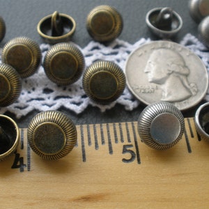 May include: A collection of 14 antique-style buttons in two finishes: bronze and silver. The buttons have a raised, circular design and are arranged on a white lace doily and a wooden ruler.