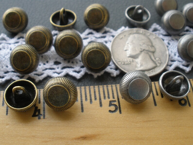 May include: A collection of 14 antique-style buttons in two finishes: bronze and silver. The buttons have a raised, circular design and are arranged on a white lace doily and a wooden ruler.