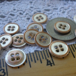 Puede incluir: Una colecci&oacute;n de 12 botones de plata y cobre con cuatro agujeros cada uno. Los botones est&aacute;n dispuestos sobre un mantel de encaje blanco y una regla de madera.