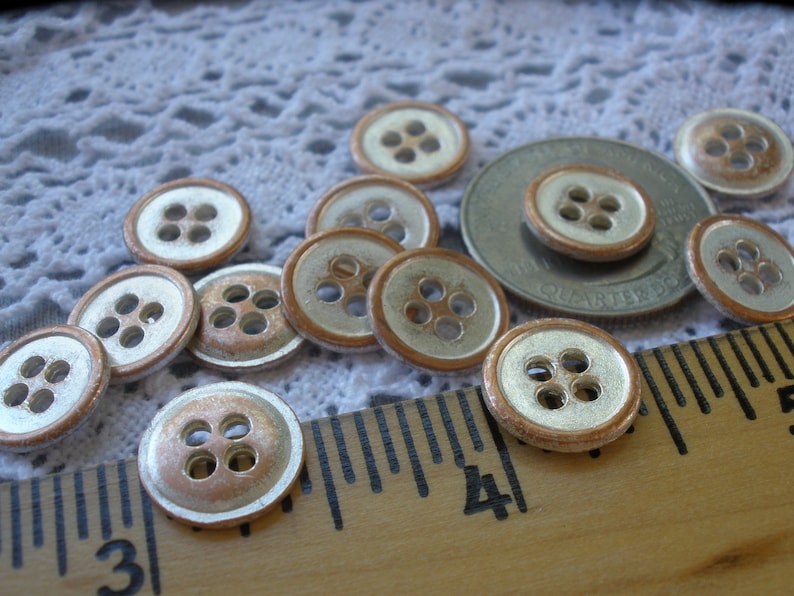 Puede incluir: Una colecci&oacute;n de 12 botones de plata y cobre con cuatro agujeros cada uno. Los botones est&aacute;n dispuestos sobre un mantel de encaje blanco y una regla de madera.