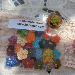 May include: A clear plastic bag containing small, multi-colored crochet flowers in 25 different shades, each with a yellow center. A white label reads 25 colors crochet flowers www.kabooco.com. A copper penny is placed inside for scale.