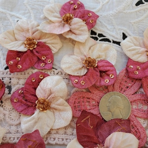 May include: A collection of fabric flower appliques in shades of cream and deep rose pink. Each flower features layered petals, a central gold-toned knot, and small gold sequins. A US quarter coin is placed next to one flower for scale.