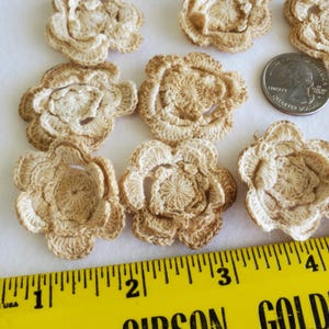 May include: A collection of handmade crochet flowers in shades of beige and light brown. The flowers are arranged on a white surface, with a yellow ruler and a quarter coin for size reference. The flowers have a detailed, textured appearance.