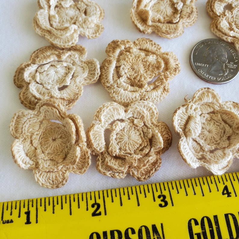 May include: A collection of handmade crochet flowers in shades of beige and light brown. The flowers are arranged on a white surface, with a yellow ruler and a quarter coin for size reference. The flowers have a detailed, textured appearance.