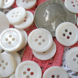 May include: A close-up of a pile of white, round, four-hole buttons. The buttons are made of a pearlescent material and have a smooth, shiny finish. A US quarter is included for scale.