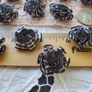May include: Several small, handmade fabric flowers with a black and white checkered pattern. The flowers are arranged on a wooden ruler and a white lace surface. A silver coin is also visible.