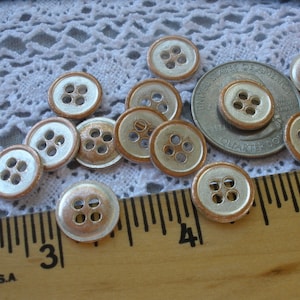 Puede incluir: Una colecci&oacute;n de 11 botones plateados y de cobre, cada uno con cuatro agujeros. Los botones est&aacute;n dispuestos sobre un mantel de encaje blanco con una regla en primer plano.