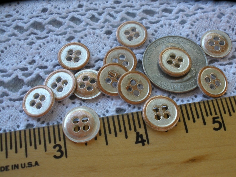 Puede incluir: Una colecci&oacute;n de 11 botones plateados y de cobre, cada uno con cuatro agujeros. Los botones est&aacute;n dispuestos sobre un mantel de encaje blanco con una regla en primer plano.