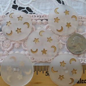 May include: A collection of five round, off-white buttons with a celestial design. Each button features gold-colored stars and crescent moons. A U.S. quarter is included for size comparison. The buttons are on a lace background.