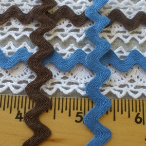 May include: Two zig-zag trim ribbons, one brown and one blue, are shown on top of a white crocheted fabric. A ruler is placed below the fabric with the numbers 4, 5, and 6 visible.