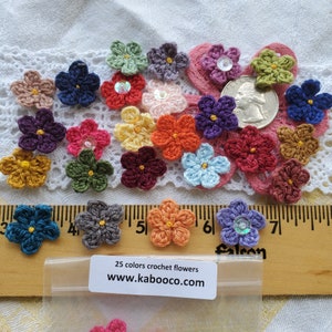 May include: A collection of 25 colorful crocheted flowers, each with a unique design and color. The flowers are arranged on a white crocheted surface and a wooden ruler. The flowers are approximately 1 inch in diameter. The text "25 colors crochet flowers www.kabooco.com" is printed on a white label.