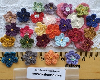 Puede incluir: Una colección de 25 flores de crochet de colores, cada una con un diseño y color únicos. Las flores están dispuestas sobre una superficie de crochet blanco y una regla de madera. Las flores tienen un diámetro de aproximadamente 2,5 cm. El texto "25 colors crochet flowers www.kabooco.com" está impreso en una etiqueta blanca.