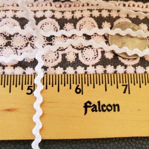 May include: White wavy trim ribbon with a scalloped edge, shown against a pink lace fabric and a wooden ruler. The ruler measures in inches and has the word "Falcon" printed on it. A US quarter is also visible.