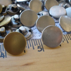 May include: A close-up of a variety of round metal buttons in gold and silver tones. The buttons are arranged on a wooden ruler with markings in inches.