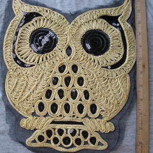May include: A gold-colored, embroidered owl applique with black sequin accents. The owl is stitched onto a gray fabric, with a black mesh backing. A wooden ruler is placed next to the applique.