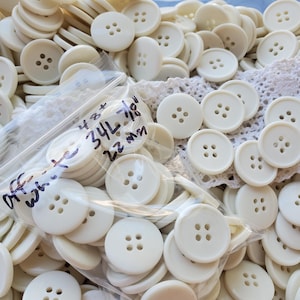 May include: A large collection of round, off-white plastic buttons, each featuring four holes in the center. A clear plastic bag contains a portion of the buttons, with handwritten text on the bag reading 48+ 34L 7/8" 22 mm.