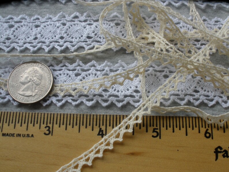 May include: Two pieces of white lace trim with a scalloped edge. The lace is laid out on a wooden ruler with the numbers 2, 3, 4, 5, and 6 visible. A silver US quarter is also visible.