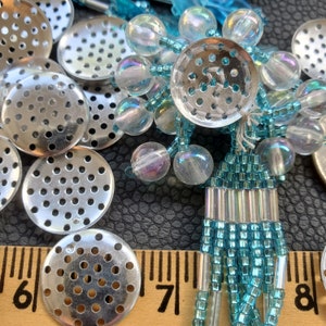 May include: A close-up of a silver metal mesh disc with a beaded tassel. The tassel is made of turquoise blue beads and silver beads. The mesh disc is surrounded by iridescent beads.