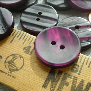 K&ouml;nnte beinhalten: Eine Sammlung von violetten und schwarzen Plastikkn&ouml;pfen mit zwei L&ouml;chern. Die Kn&ouml;pfe sind auf einem Holzlineal mit den Worten "Shoes" und "New" darauf angeordnet.