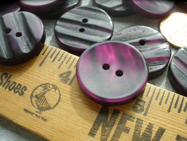 K&ouml;nnte beinhalten: Eine Sammlung von violetten und schwarzen Plastikkn&ouml;pfen mit zwei L&ouml;chern. Die Kn&ouml;pfe sind auf einem Holzlineal mit den Worten "Shoes" und "New" darauf angeordnet.
