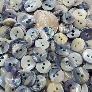 May include: A large pile of two-hole iridescent shell buttons in shades of white, gray, and purple. The buttons are oval shaped and have a smooth, shiny finish.