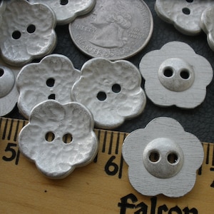 May include: A collection of silver-colored metal flower-shaped buttons with two holes each. The buttons have a textured surface and are arranged on a wooden surface with a ruler.