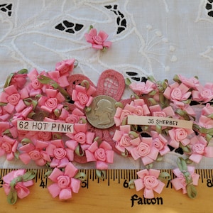 May include: A close-up of a collection of pink satin ribbon flowers. The flowers are arranged on a white lace fabric background. The flowers are labeled with the numbers 62 and 35, and the words "Hot Pink" and "Sherbet".