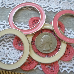 May include: Close-up of several decorative rings with a silver rim and a pink or cream-colored enamel finish. A U.S. quarter is centered within a pink crocheted flower. The rings are arranged on a gray surface with white lace.