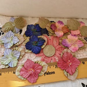 May include: A collection of fabric flower appliqués in shades of blue, pink, and purple, each featuring green leaves and a sequin center. A US quarter coin and a wooden ruler showing inches are placed among the flowers for scale reference.