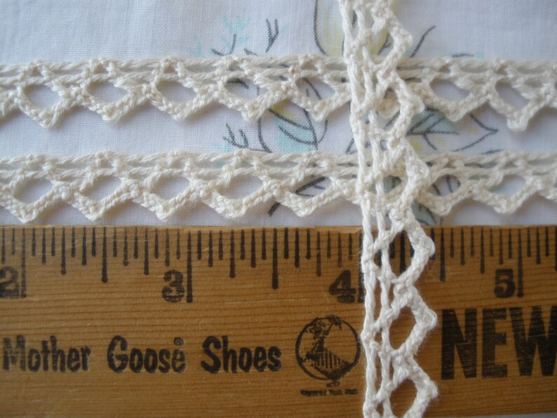 May include: White crochet trim with a delicate diamond pattern. The trim is measured against a wooden ruler with the words "Mother Goose Shoes" printed on it.