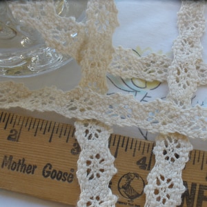 May include: Close-up of a white crocheted lace trim with a scalloped edge. The lace is laid out on a white surface with a ruler underneath. The ruler has the words "Mother Goose" printed on it.