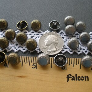 May include: A collection of 18 round metal buttons in two colors: antique gold and silver. The buttons have a raised rim and a flat center. The buttons are arranged on a white lace doily and a wooden ruler.