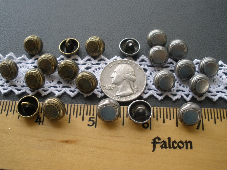 May include: A collection of 18 round metal buttons in two colors: antique gold and silver. The buttons have a raised rim and a flat center. The buttons are arranged on a white lace doily and a wooden ruler.