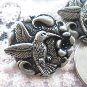 May include: Two silver-toned metal buttons featuring a hummingbird in a floral design. The buttons are round and have a detailed, textured surface.