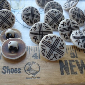 May include: A collection of vintage buttons with a cream-colored base and dark brown crosshatch and starburst designs. The buttons are round and have a shank on the back. A wooden ruler with the text "NEW T" is in the foreground.