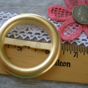 May include: A gold-colored metal belt buckle with a round, smooth, ring-shaped design. The buckle is approximately 2 inches in diameter and has a metal bar that slides through the ring.