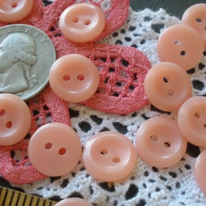 Può includere: Una collezione di bottoni di plastica rosa con due fori ciascuno. I bottoni sono disposti su un pizzo bianco all'uncinetto con un pizzo rosa all'uncinetto sotto.