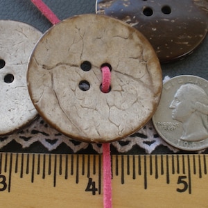 May include: Three round, brown, coconut shell buttons with a textured surface. One button has three holes and is tied with a pink string. The other two buttons have four holes. A ruler is in the background.