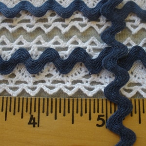 May include: Blue zig zag trim with a wavy edge. The trim is shown on a white lace fabric and a ruler marked in inches.