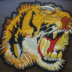 May include: Embroidered patch featuring a roaring tiger with yellow, orange, and black details. The tiger's mouth is open, revealing red and white teeth. The patch has a black border and is likely for jackets or bags.